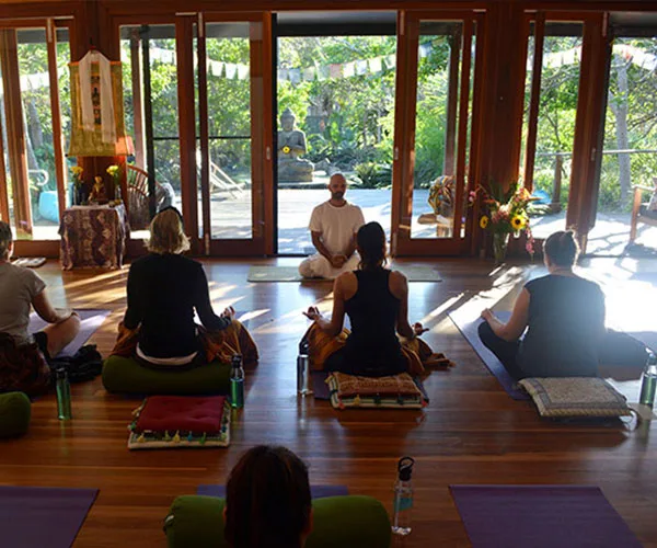 Group meditation session indoors with an instructor, surrounded by nature through glass doors.