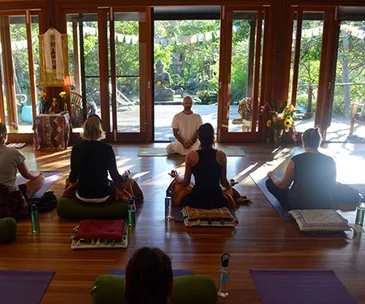 Group meditation session indoors with an instructor, surrounded by nature through glass doors.