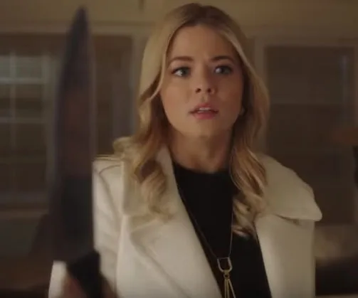 Woman with long blonde hair in a white coat looks surprised, holding a large knife in a dimly lit room.