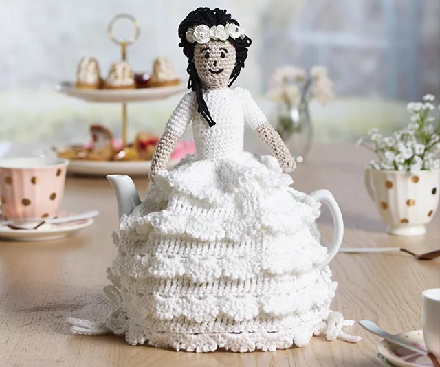 Crocheted tea cosy resembling a princess in a white dress with black hair and a flower crown, on a table set for tea.