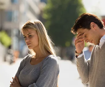 A man and woman are standing outdoors, both appearing upset and thoughtful, with the man rubbing his forehead.