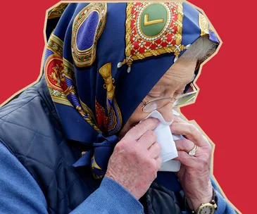 An elderly person wearing a colorful headscarf cleans glasses with a cloth against a red background.