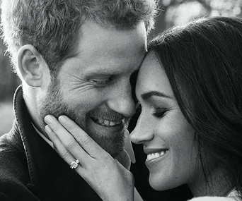 A couple smiling lovingly, posing closely with the woman's hand showing an engagement ring on the man's cheek in black and white.