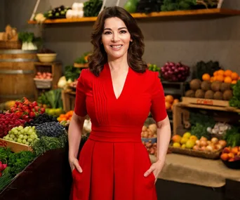 Woman in a red dress stands in front of an array of fresh fruits and vegetables in a market setting.