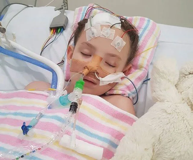 A child lies in a hospital bed with medical tubes and electrodes, covered by a colorful blanket.