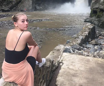 Young woman sitting by a rocky riverbank, looking back with a waterfall in the background.