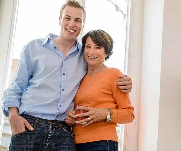 A young man in a light blue shirt with an older woman in an orange sweater, smiling and standing together by a window.