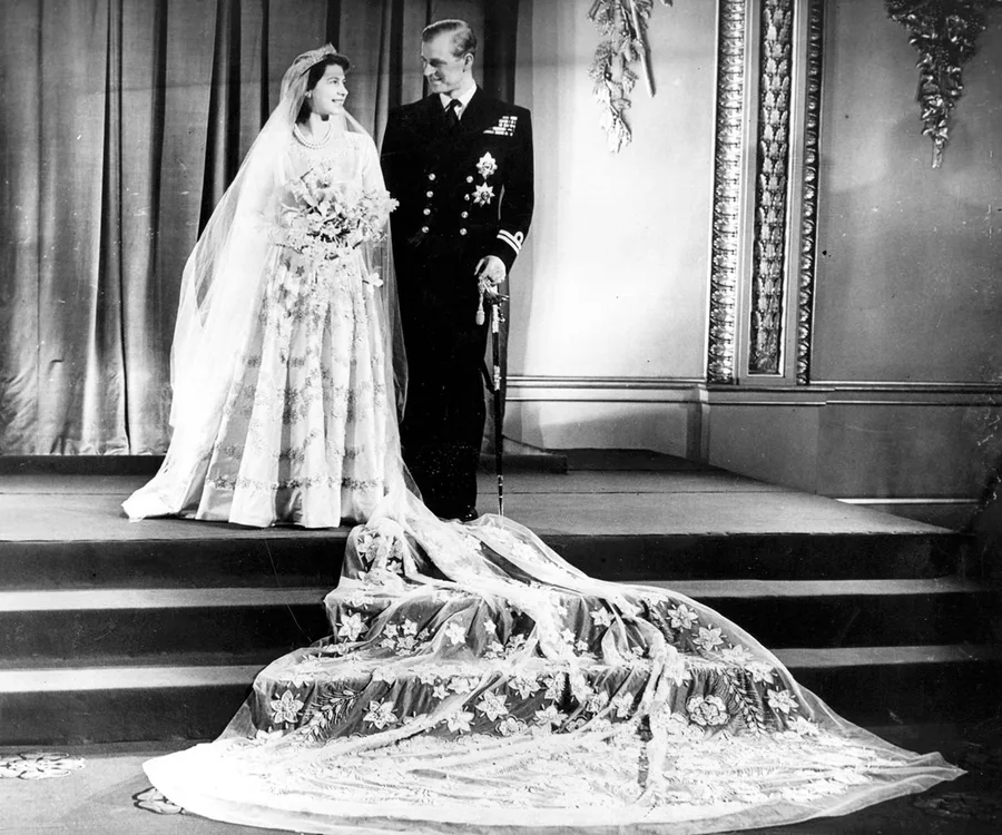 A bride and groom stand on a platform, smiling at each other. The bride's train is detailed with floral patterns.