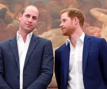 Two men in suits converse in front of a painting in a formal setting.