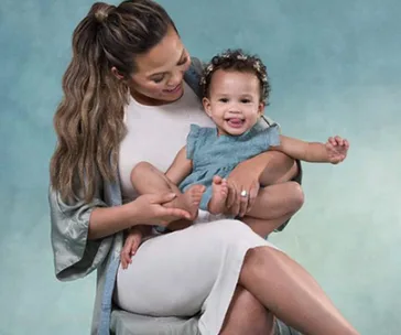 Woman smiling while holding a happy baby on her lap against a soft, blue background.