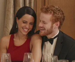 A smiling couple in formal attire seated at a dining table, sharing a light moment at an event.