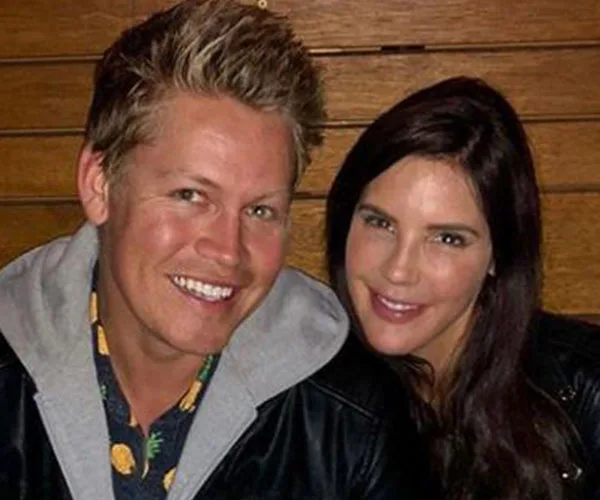 A smiling man and woman sitting closely, wearing casual jackets, with wooden background.