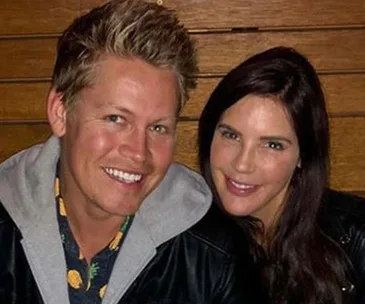 A smiling man and woman sitting closely, wearing casual jackets, with wooden background.