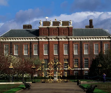 Kensington Palace's front view with ornate golden gates and a red-brick facade under a blue sky with clouds.