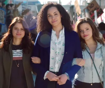 Three women smile and link arms while walking in a protest with signs visible in the background.