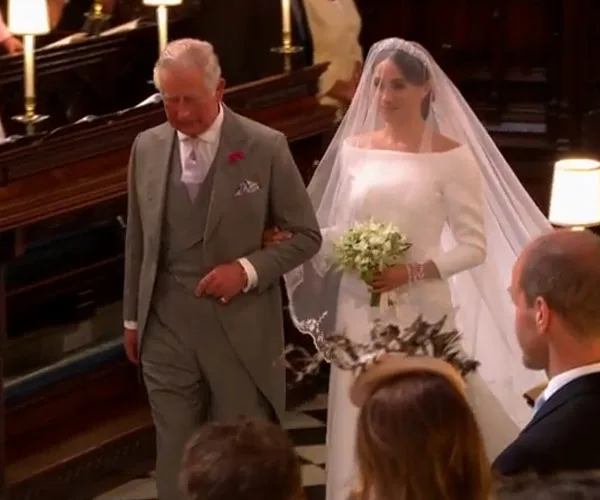 Prince Charles walks Meghan Markle down the aisle in a wedding setting, surrounded by guests.