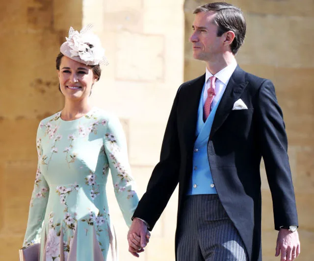 A woman in a floral dress and fascinator holds hands with a man in morning attire at an outdoor event.