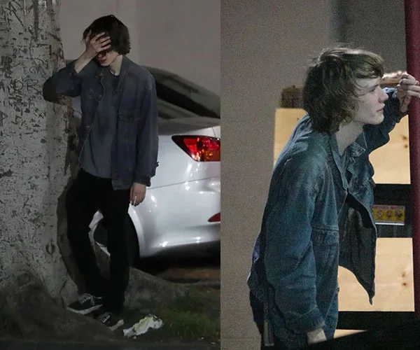 Young person in denim jacket outside, leaning on a tree and a structure, appears contemplative or weary.