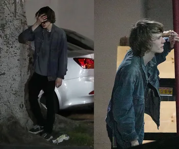 Young person in denim jacket outside, leaning on a tree and a structure, appears contemplative or weary.