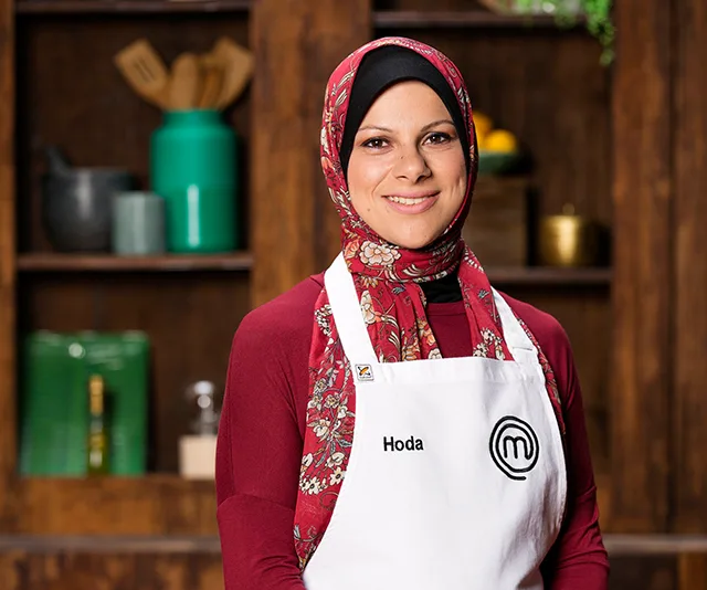 A woman smiling, wearing a red floral hijab and a white apron with "Hoda" and a MasterChef logo.