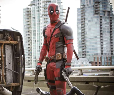 "Deadpool in a red and black costume with guns, standing on a city street with skyscrapers in the background."