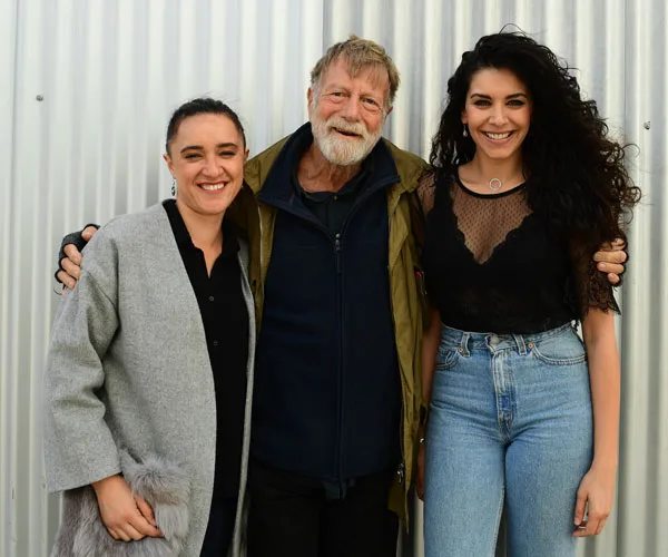 Group photo of three smiling people, two women and a man, standing in front of a corrugated metal background.