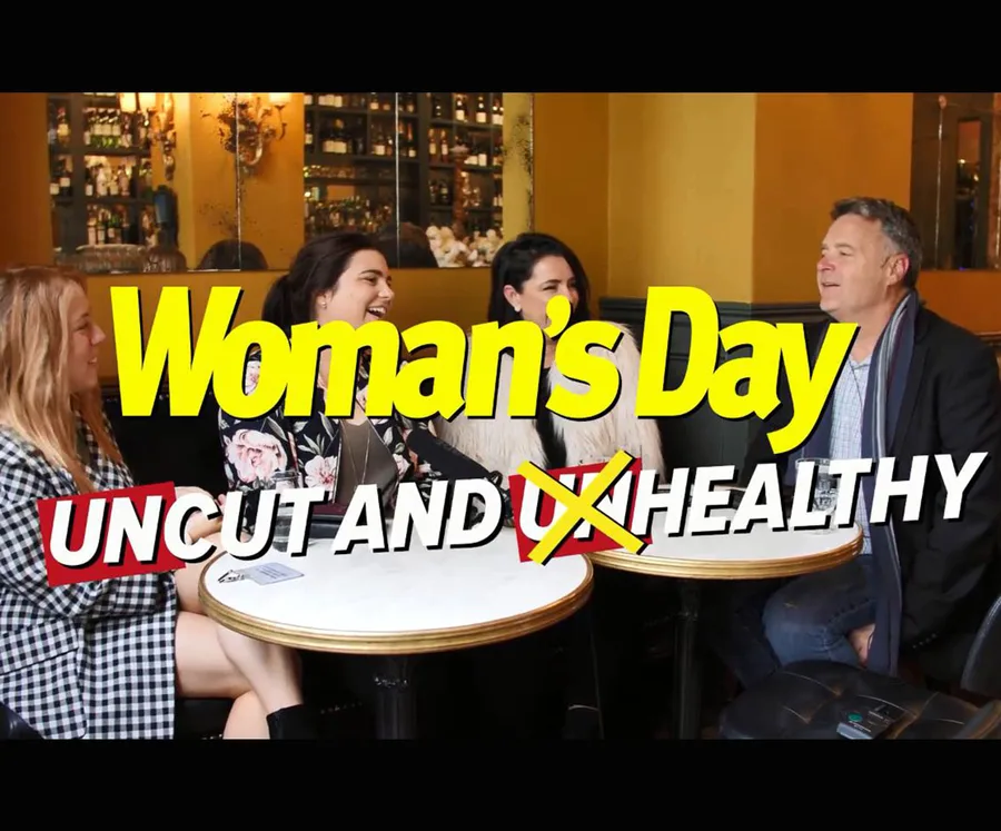 Group of people laughing at a table in a cafe with "Woman's Day" and "Uncut and Healthy" text overlay.