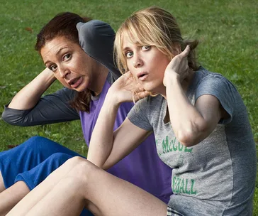 Two women doing sit-ups on grass, wearing casual workout clothes, looking surprised or concerned.