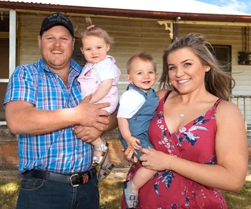 Family of four smiling outdoors, standing in front of a house. Parents hold two young children.