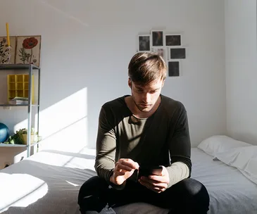 Man sitting on a bed using a smartphone in a sunlit room with wall art and shelves in the background.