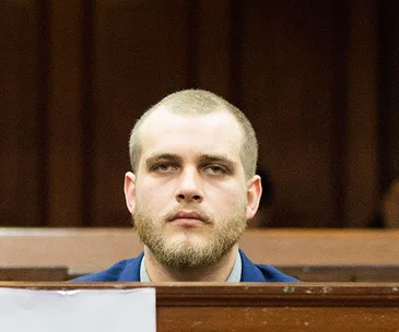 Man sitting in a courtroom, wearing a blue suit, looking forward.