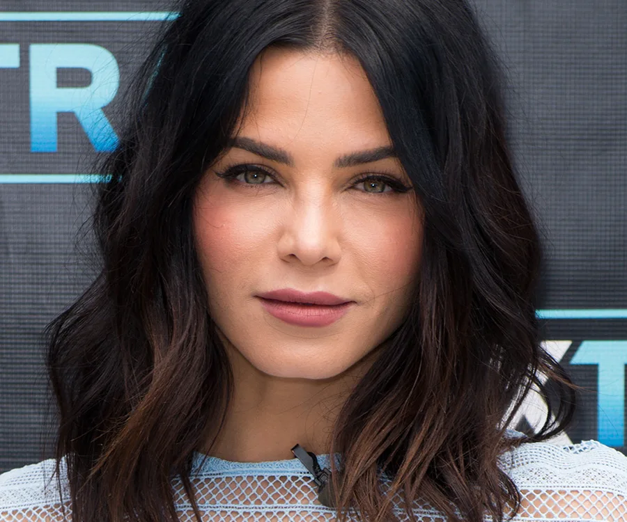 A woman with shoulder-length dark hair, wearing a lace top against a media backdrop.