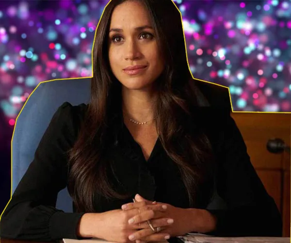 Woman with long hair wearing black, sitting at a desk with colorful bokeh in the background.