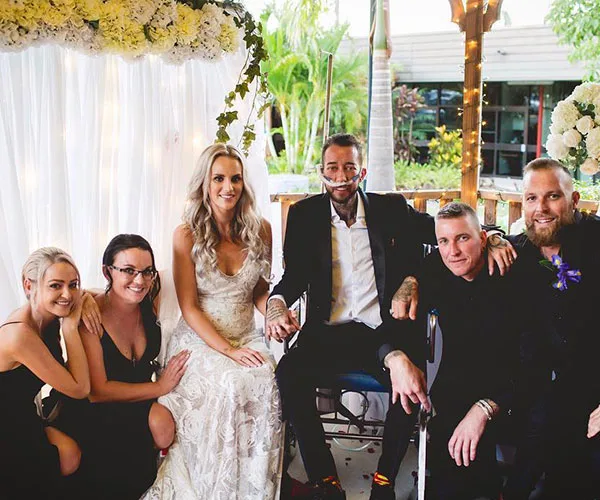 Bride and terminally ill groom in a wheelchair at their wedding, surrounded by friends, under a floral arch.
