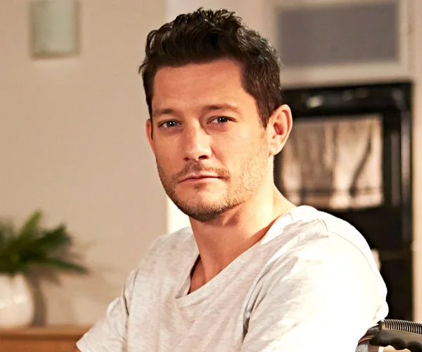 Finn Kelly seated indoors, wearing a white shirt, with a neutral expression.