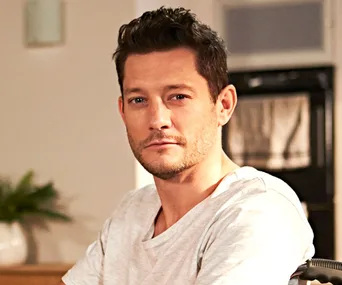 Finn Kelly seated indoors, wearing a white shirt, with a neutral expression.
