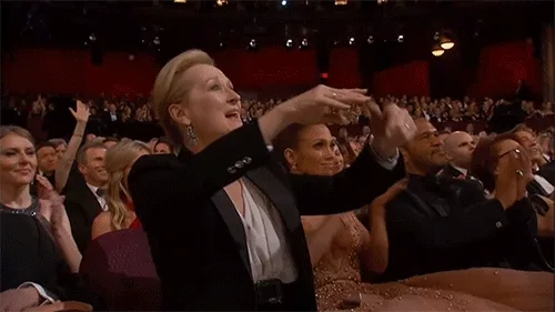 A woman in an elegant suit enthusiastically clapping and cheering in a crowded theater.