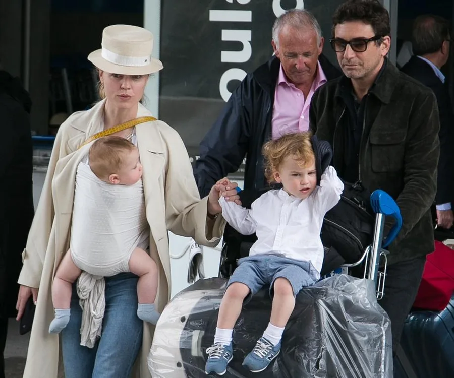 Family traveling with luggage and two children; one baby in a carrier, another child on a luggage cart.