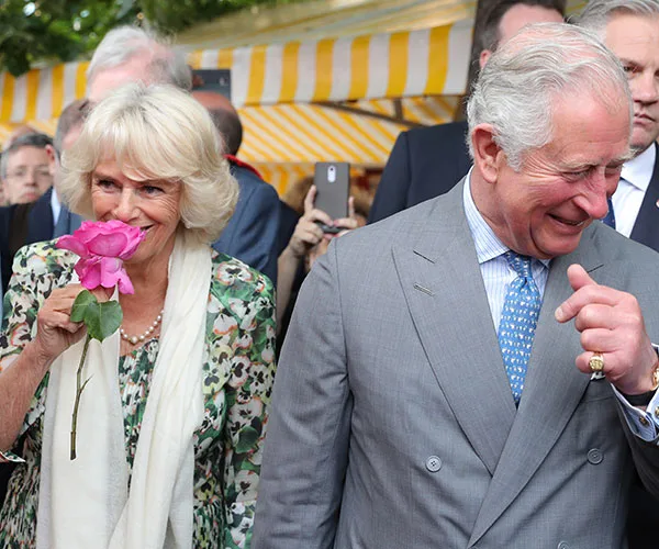 Camilla and Charles smiling outdoors, Camilla holding a pink rose, with a striped tent in the background.