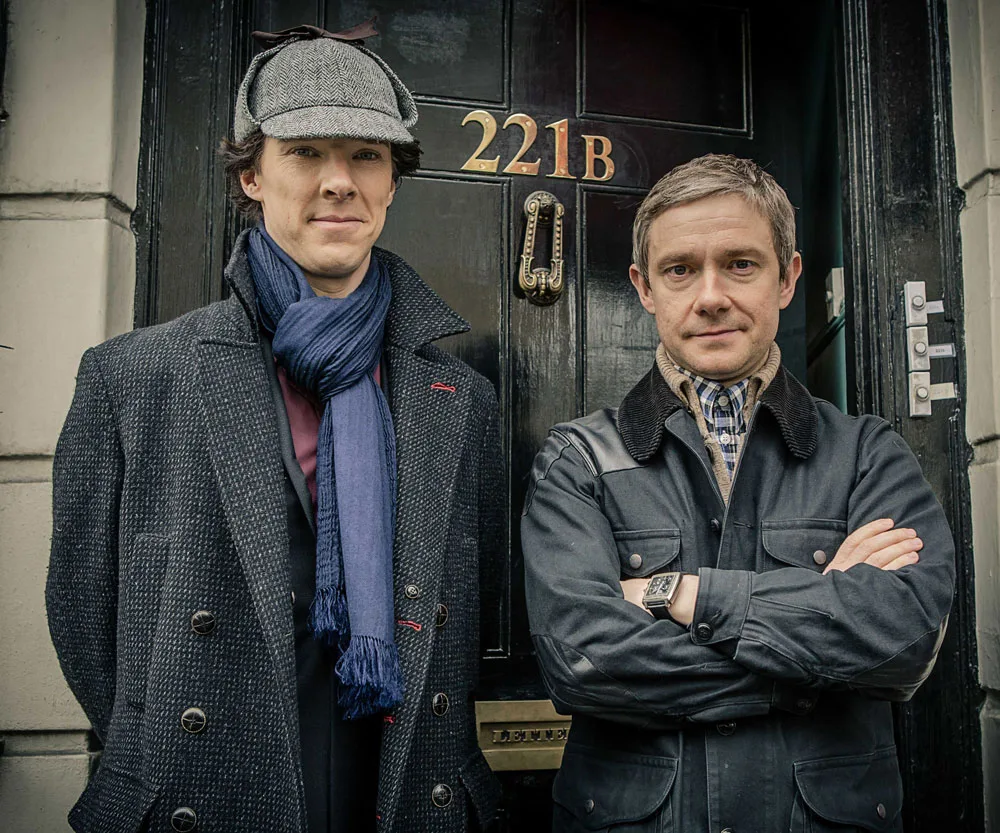 Two men at 221B Baker Street door, one in a deerstalker hat and scarf, the other in a jacket, portraying Sherlock Holmes and Watson.