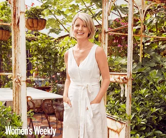 A woman in a white dress smiles, standing in a lush garden with a rustic metal archway.