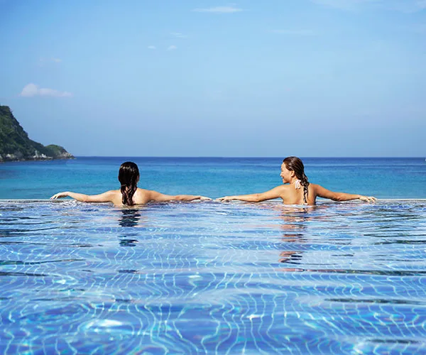 Two women relaxing in an infinity pool with a view of the ocean and a distant hillside under a clear blue sky.