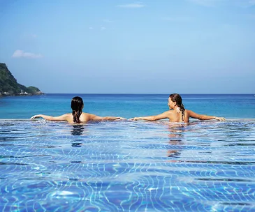 Two women relaxing in an infinity pool with a view of the ocean and a distant hillside under a clear blue sky.