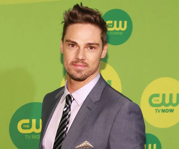 Jay Ryan at a CW event, wearing a suit with a striped tie, standing in front of a green background with CW logos.