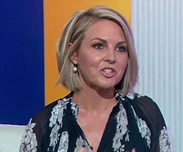 A woman with short blonde hair and pearl earrings speaks on a TV set with a colorful backdrop.
