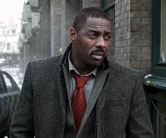 Idris Elba in a dark coat and red tie, walking on a snowy street, looking serious.