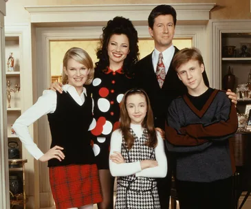 "The Nanny cast in a living room; five characters, including a woman in a polka dot dress, smile for the camera."