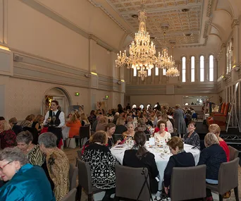 Elegant banquet hall with chandeliers, crowded tables with people dining, and waiter serving drinks.