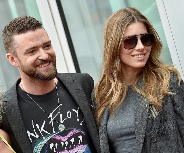 A smiling couple, the man with a beard wearing a printed shirt, and the woman with long hair wearing sunglasses and a gray jacket.