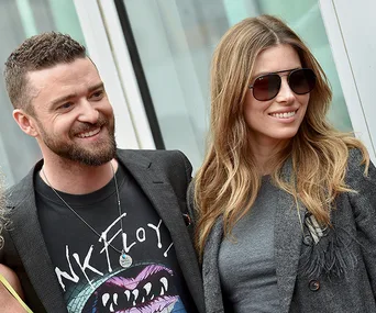 A smiling couple, the man with a beard wearing a printed shirt, and the woman with long hair wearing sunglasses and a gray jacket.